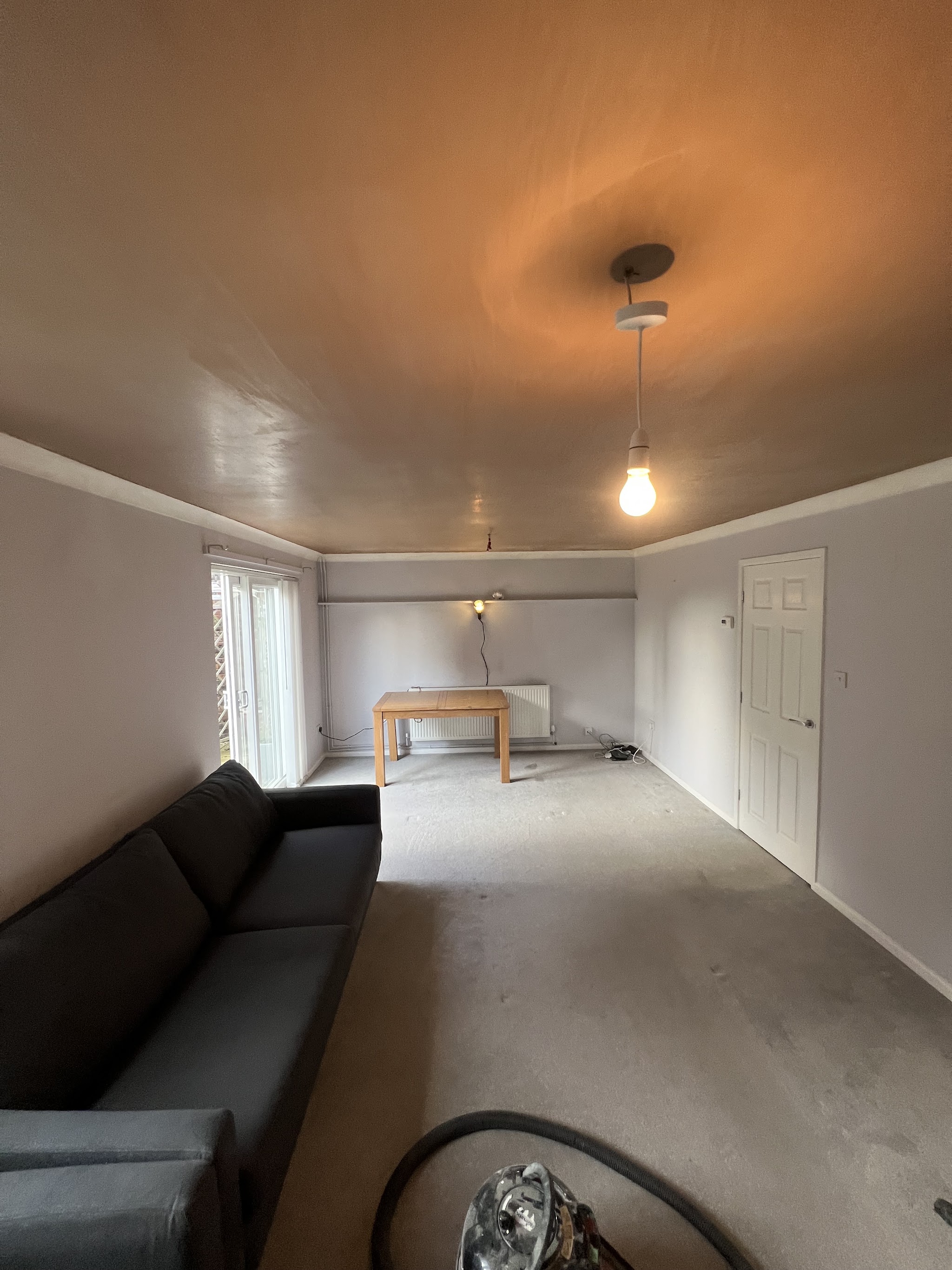 A long living room with the walls and ceiling freshly plastered, a sofa and a pendant light